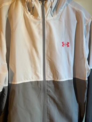  Under Armour jacka  - Snygg vindjacka från Under Armour i grått och vitt med dragkedja framtill och huva. Jackan har meshfoder och en rosa logga på bröstet. Perfekt för blåsiga dagar och sportiga outfits. Använd 1-2 gånger max 