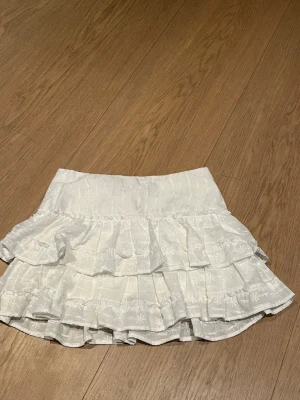 Vit volangkjol från H&M - Supersöt vit kortkjol från H&M med volanger och broderade blommor. Kjolen har en bred midja och flera lager volanger som ger en luftig och lekfull look. Perfekt att styla med en enkel topp för en fräsch outfit.