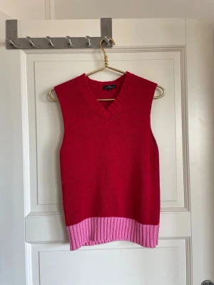 Röd stickad väst från Day Birger - Snygg röd stickad väst från Day Birger et Mikkelsen med v-ringad hals och bred rosa ribbad kant nedtill. Perfekt att styla över en skjorta eller t-shirt för en trendig lager-på-lager-look.