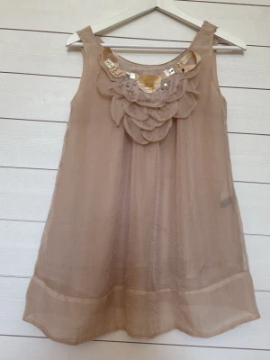 Beige ärmlös kortklänning med volang - Beige ärmlös kortklänning med volangdetaljer och pärlor framtill. Klänningen har en flowy och luftig passform, perfekt för varma sommardagar. Tillverkad i ett tunt material som ger en lätt känsla.