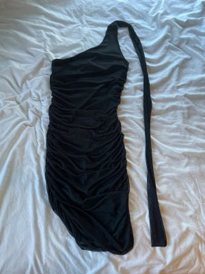Svart one shoulder klänning - Snygg svart kortklänning med one shoulder-design och rynkade detaljer. Klänningen har en lång axelband som kan knytas och är tillverkad i ett mjukt, stretchigt material. Perfekt för dig som vill sticka ut med en trendig look.