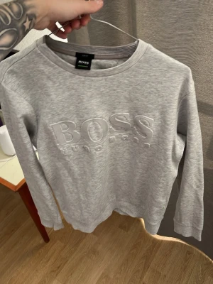 Grå sweatshirt från Hugo Boss - Snygg ljusgrå sweatshirt från Hugo Boss med stort broderat BOSS-tryck på bröstet. Tröjan har rund halsringning, långa ärmar och ribbade muddar vid ärmslut och nederkant. Tillverkad i mjukt bomullsmaterial för en skön och avslappnad look.