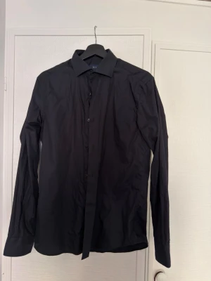 Svart slim fit skjorta från Riley - Säljer en svart skjorta från Riley med slim fit-passform. Skjortan har klassisk krage, knappar framtill och långa ärmar. Perfekt för dig som gillar en stilren look. Materialet känns lätt och bekvämt. 