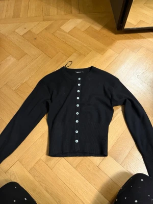 Svart kofta från Gina Tricot - Stilren svart kofta från Gina Tricot med rund halsringning och långa ärmar. Koftan har ribbade muddar och knappar hela vägen framtill i kontrasterande färg. Perfekt att matcha med jeans eller kjol för en enkel och snygg look.