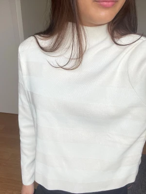Vit ribbad tröja med hög krage - Snygg vit långärmad tröja med ribbad struktur och hög krage. Tröjan har en rak passform och är tillverkad i ett mjukt, stretchigt material som känns skönt mot huden. Perfekt att styla med jeans eller kjol för en clean look.