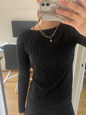Svart plisserad långärmad topp - Säljer en svart långärmad topp med plisserad struktur och rund halsringning. Toppen har en slim passform och är tillverkad i ett mjukt syntetmaterial som känns bekvämt mot huden. Perfekt att matcha med jeans eller kjol för en stilren look.