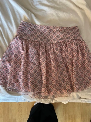 Mönstrad kortkjol från Meet Me There - Supersöt kortkjol från Meet Me There med blommigt mönster i rosa, rött och blått. Kjolen har en bred linning och ett lätt, fladdrigt tyg som ger en luftig känsla. Den har shorts under.