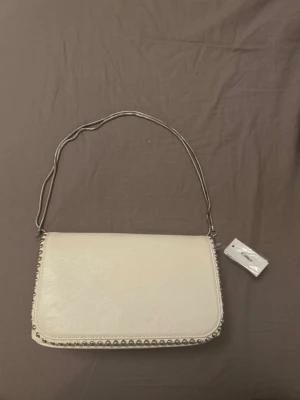 Vit handväska med silverdetaljer - Jättefin vit handväska från Glitter som är helt oanvänd. Väskan har silverfärgad kedjerem och dekorativa nitar längs kanterna. Väskan har ett ”lock” med magnetknäppning och är tillverkad i ett slätt, konstläderliknande material.                    Nypris: 299kr                                                             Pris: 100kr
