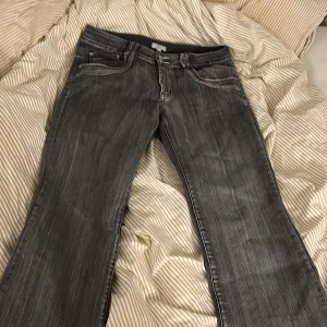 Grå bootcut jeans med slitningar - Säljer ett par grå bootcut jeans med diskreta slitningar och klassiska femficksdetaljer. Jeansen har en låg midja och är tillverkade i denim med stretch. Perfekta för dig som gillar en avslappnad och trendig look. Storlek står 44 men passar storlek 38-40. Köptes second hand och är uppsydda nedtill men passar mig som är 168