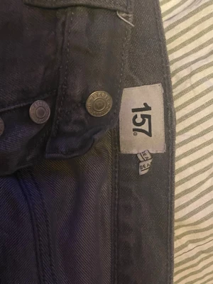 Gråa jeans från 157 - Säljer ett par mörk grå jeans från 157 med klassisk femficksdesign och metallknappar. Jeansen har en smal passform och är tillverkade i denim. Perfekt för dig som gillar stilrena och tidlösa jeans.