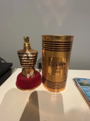 Jean Paul Gaultier Le Male Elixir - Lyxig parfym i metallisk guldflaska formad som en manlig torso, med matchande guldfärgad cylinderförpackning och röd sammetsbotten. Flaskan har eleganta ränder och en exklusiv känsla. Perfekt för dig som vill sticka ut med en ikonisk doft.