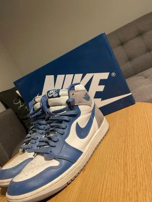 Nike Air Jordan 1 High blå/vit - Säljer ett par Nike Air Jordan 1 High sneakers i blått och vitt med grå detaljer. Skorna har klassisk hög siluett, Nike Swoosh på sidan och är tillverkade i läder med textildetaljer. Perfekta för dig som gillar streetwear och ikonisk design.