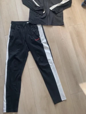 Nike träningsset - Snyggt grått träningsset från Nike med jacka och byxor. Jackan har huva, dragkedja och två fickor, medan byxorna har resår i midjan och vid benslut. Båda plaggen har Nike-logga i vitt. Perfekt för träning eller sportiga dagar. Storleken är XS, sitter som S. Passar mellan 175-185 cm