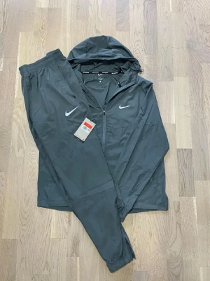 Nike svart träningsset grå - Snyggt svart träningsset från Nike med vita ränder längs ärmar och ben. Byxorna har dragsko i midjan och röd Nike-logga på låret. Perfekt för träning eller chill. Materialet är mjukt och stretchigt för maximal komfort.
