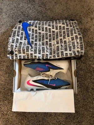 Nike Phantom Elite fotbollsskor - Nike Phantom Elite fotbollsskor i vitt, blått och svart med rosa detaljer. Skorna har snörning, mönstrad ovandel för bättre bollkontroll och en sula med dobbar för optimalt grepp på planen. Levereras med originalpåse och låda. 