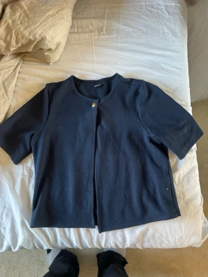 Mörkblå kofta från Gina Tricot - Mörkblå kofta från Gina Tricot med kort ärm och rund halsringning. Stängs med en silvrig knapp upptill och har en rak, enkel passform. Perfekt för lager-på-lager och stilrena outfits.