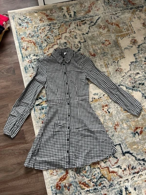 Svartvit rutig skjortklänning - Snygg svartvit rutig skjortklänning med lång ärm och knappar hela vägen framtill. Klänningen har klassisk krage och markerad midja som ger en feminin siluett. Perfekt för dig som gillar en tidlös och trendig look.