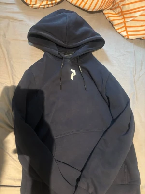 Mörkblå hoodie från Peak Performance - Snygg mörkblå hoodie från Peak Performance med klassisk huva och dragsnören. Framtill finns en vit logga och en stor magficka. Perfekt för en avslappnad och sportig stil. Mjuk bomullsblandning som känns skön mot huden.