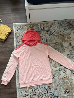 Rosa hoodie med tryck och ränder - Snygg hoodie i ljusrosa med korallröd huva och axelparti. Tryck på bröstet med texten 'HAPPINESS noun a giggle wrapped in sunshine & glitter'. Vita ränder längs ärmarna. Perfekt för en avslappnad och trendig stil.