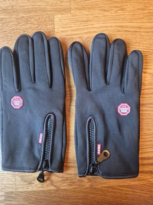 Svarta sportiga handskar med dragkedja - Svarta handskar från Kyncilor med dragkedja och greppvänlig insida. Perfekta för kyliga dagar och har en sportig look. Touchfunktion på fingertopparna och diskret logga på ovansidan. Snygga och praktiska accessoarer för uteliv.