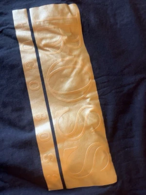 Svart t-shirt med guldtryck från BOSS - Svart t-shirt från BOSS med stort guldtryck och logga framtill. Klassisk rund hals och korta ärmar. Snygg och stilren design som sticker ut med den metalliska detaljen. Perfekt för dig som gillar märkeskläder och vill ha något extra.