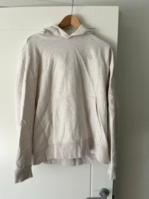 Beige hoodie från Calvin Klein - Säljer en beige hoodie från Calvin Klein med broderad CK-logga på bröstet. Hoodien har en stor huva, ribbade muddar och en klassisk känguruficka framtill. Materialet är mjukt och bekvämt, perfekt för chill dagar. Passar dig som gillar stilren design.priset kan diskuteras 