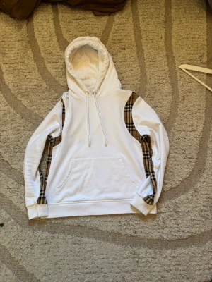 Burberry hoodie  - Vit hoodie från Burberry i storlek S, inköpt för 6000 kr. Tröjan är i mycket gott skick utan några större defekter – endast en liten, knappt märkbar fläck finns. Klassisk Burberry-design med de ikoniska rutiga panelerna på sidorna som ger en exklusiv och stilren look. Perfekt för både vardag och en mer uppklädd casual stil.