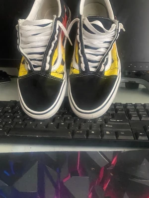 Vans Old Skool med flame-mönster - Säljer ett par Vans Old Skool sneakers med svart tå och häl, vit sula och snörning samt ett färgstarkt flame-mönster i gult, orange och rött på sidorna. Klassisk vit sidestripe och canvas/suede-material. Perfekta för dig som vill sticka ut med din stil.