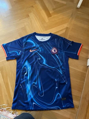 Chelsea FC Nike matchtröja blå - Chelsea FC matchtröja från Nike i blått med unikt mönster i ljusblått och vitt. Tröjan har korta ärmar, rund hals och klubbmärke på bröstet. Orange Nike-logga och röda detaljer vid ärmslut. Perfekt för fotboll eller träning.