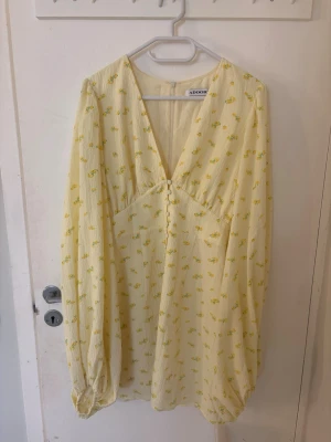 Ljusgul blommig klänning från Adoore - Supersöt ljusgul klänning från Adoore med små gula blommor, v-ringning och långa ärmar. Klänningen har knappar framtill och ett lätt, luftigt tyg som passar perfekt till varmare dagar.