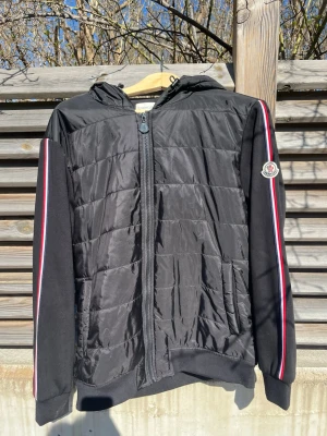 Svart pufferjacka från Moncler - Snygg svart pufferjacka från Moncler med huva och dragkedja framtill. Jackan har quiltad front, ribbade muddar och röda, vita och blå ränder längs ärmarna. Moncler-logga på vänster ärm. Perfekt för kalla dagar och riktigt stilren.