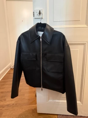 Svart jacka från Carin Wester - Cool svart jacka i skinnimitation från Carin Wester med rak passform, stora bröstfickor och bred krage. Jackan har en kraftig dragkedja framtill. Perfekt för dig som vill ha en stilren och modern look.