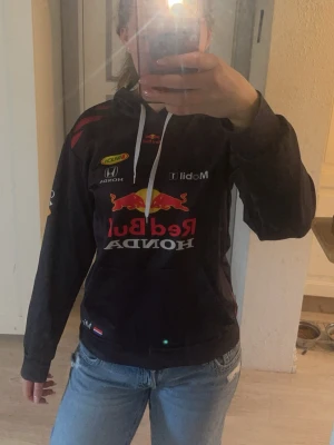 Red Bull Racing hoodie med tryck - Svart hoodie från Red Bull Racing med stora tryck av Red Bull, Honda och Mobil 1 på både fram- och baksidan. Hoodien har huva med vita snören och känguruficka framtill. Säljer för den inte används💜Kolla in min profil för fler fina och trendiga plagg!