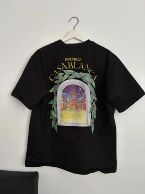Svart t-shirt från Casablanca - Cool svart t-shirt från Casablanca med stor färgglad grafik på ryggen som föreställer en byggnad och texten 'Avenida Casablanca'. T-shirten har korta ärmar och är gjord i mjuk bomull. Perfekt för dig som gillar unika prints och streetstyle.