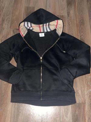Burberry hoodie - Snygg svart hoodie från Burberry med dragkedja framtill och klassiskt rutmönster i huvan. Hoodien har fickor på sidorna och ribbade muddar vid ärmslut och nederkant. Perfekt för dig som vill ha en stilren men ändå unik look.