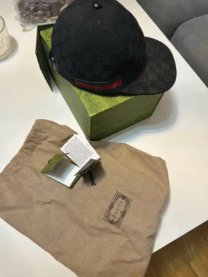 Svart keps med monogram - Snygg svart keps från  med klassiskt monogram över hela ytan och en grön-röd rand på sidan. Kepsen har böjd skärm och justerbar rem baktill.