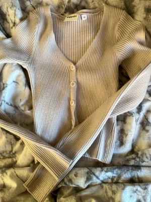 Beige ribbad kofta från Kappahl - Snygg beige ribbstickad kofta från Kappahl med v-ringning och tre knappar framtill. Långa ärmar och figurnära passform gör den perfekt att styla till jeans eller kjol. Enkel och stilren design som passar till många outfits.