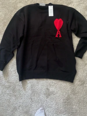 Svart stickad tröja från Ami - Svart stickad tröja från Ami Alexandre Mattiussi med rund halsringning och ribbade muddar. På bröstet finns ett rött hjärta med bokstaven A, vilket är varumärkets ikoniska detalj. Perfekt för dig som gillar stilrena och trendiga plagg.