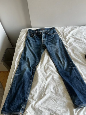 Blå jeans från Nudie Jeans - Säljer ett par klassiska blå jeans från Nudie Jeans med snygga slitningar och kontrastsömmar. Jeansen har fem fickor, raka ben och en cool läderpatch bak. Materialet är bomull och passformen är slim fit. Perfekt för dig som gillar en avslappnad och trendig stil.