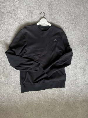Svart sweatshirt från J.Lindeberg - En stilren svart sweatshirt från J.Lindeberg med rund hals och diskret logotyp på bröstet. Tröjan har långa ärmar och ribbade muddar vid ärmslut och nederkant. Perfekt för dig som gillar enkel och klassisk design.