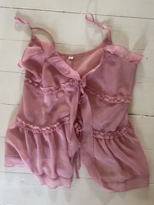 Rosa volang topp med knytband - Söt rosa topp med tunna axelband, volangdetaljer och knyt framtill. Luftigt material och lätt transparent look ger en romantisk vibe. Perfekt att styla med jeans eller kjol för en trendig outfit.