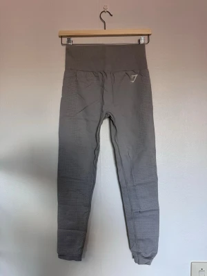 Ljusgråa seamless leggings från Gymshark - Snygga grå seamless leggings från Gymshark med hög midja och diskret mönster längs benen. Materialet är stretchigt och följsamt, perfekt för träning eller chill. Gymshark-loggan finns på höften och vaden. Riktigt bekväma och stilrena.