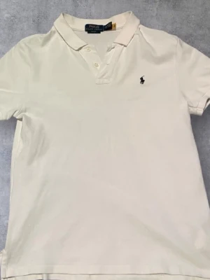 Vit polo från Ralph Lauren - Säljer en klassisk vit polo från Ralph Lauren med broderad logga på bröstet. Skjortan har korta ärmar, krage och knappar framtill. Perfekt för dig som gillar stilrena plagg och vill ha något som funkar till många outfits.