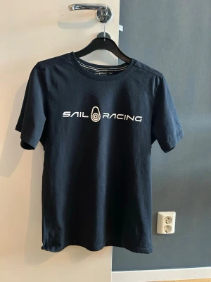 Svart Sail Racing t-shirt - Mörkblå t-shirt från Sail Racing med vit logotyp på bröstet och en mindre logga på ryggen. Klassisk rund hals och korta ärmar. Materialet mjuk bomull 