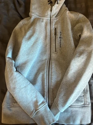 Ljusgrå hoodie från Sail Racing - Snygg ljusgrå hoodie från Sail Racing med dragkedja framtill och en bröstficka med dragkedja. Märkets logga är tryckt på bröstet. Hoodien har huva med dragsko och ribbade muddar vid ärmar och nederkant. Tillverkad i mjukt bomullsmaterial.