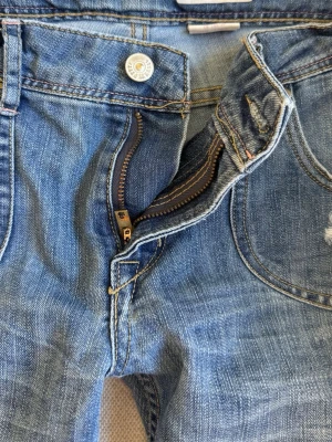Blå slitna jeans med hål - Säljer ett par blå jeans med slitningar och hål på låret och vid fickan. Jeansen har klassisk femficksmodell och bootcut och lågmidjade .