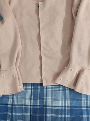 Ljusrosa blus med pärldetaljer - Supersöt ljusrosa blus med långa ärmar och volang vid ärmsluten. Ärmsluten är dekorerade med små pärlor för en extra stylish touch. Perfekt att matcha med jeans eller kjol för en trendig look.