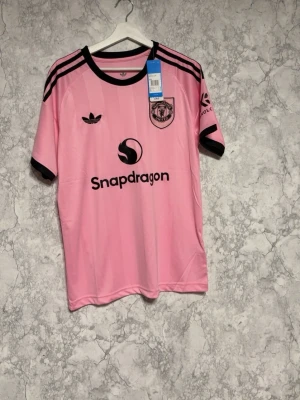 Rosa Manchester United tröja Adidas - Snygg rosa Manchester United matchtröja från Adidas med svarta detaljer och klubbmärke på bröstet. Tröjan har svarta ränder på axlarna, svart krage och är tillverkad i ett lätt och ventilerande material. Perfekt för fotbollsträning eller att bära till vardags.