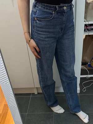 Blå raka jeans med hög midja - Säljer ett par klassiska blå jeans med rak passform och hög midja. Jeansen har fem fickor, gylf med dragkedja och knapp samt kontrastsömmar. Perfekta för en avslappnad och trendig look.