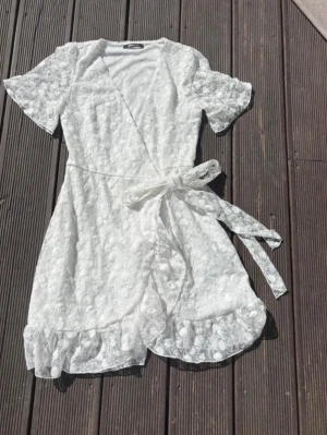 Vit spets  kläning  med volang - Superfin vit  kläning  i spets med omlottmodell och knytband i midjan. Korta ärmar och volangdetaljer längs benen ger en romantisk vibe. Perfekt för sommardagar när du vill känna dig extra söt och bekväm.